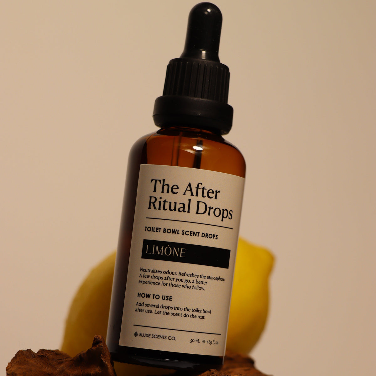 A bottle of 'The After Ritual Drops' in the 'Limone' scent, displayed on a piece of dark wood with a yellow citrus slice behind it against a plain beige background.