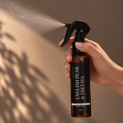A hand holding a spray bottle of English Pear & Freesia room spray, which is spraying a fine mist. The bottle is amber brown with a black spray head and features a black label with white text.