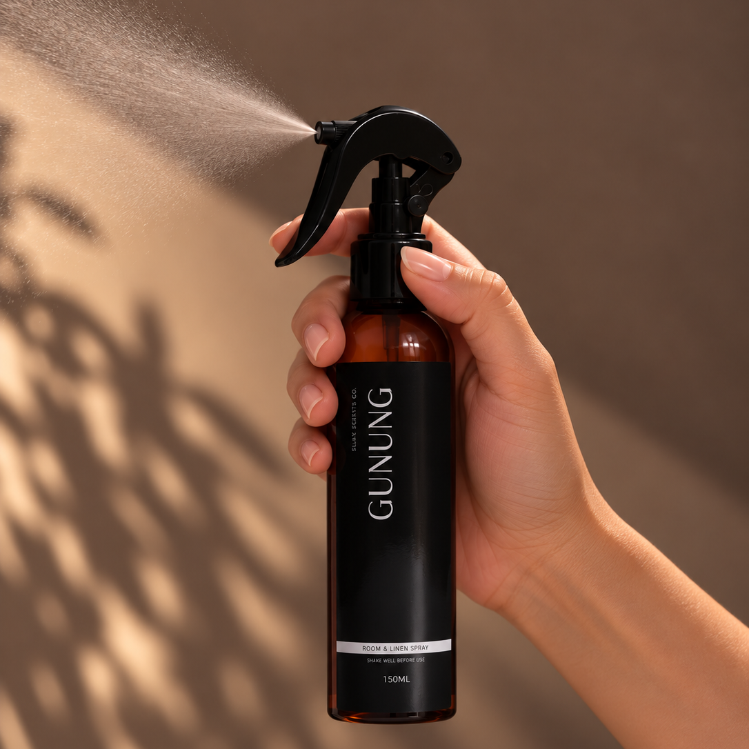 A hand holding a GUNUNG brand amber spray bottle that is spraying a fine mist, set against a warm brown background with soft leaf shadows.