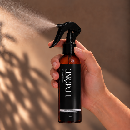 A hand holding a Limone room spray bottle that is spraying a fine mist, set against a warm brown background with leaf shadows.