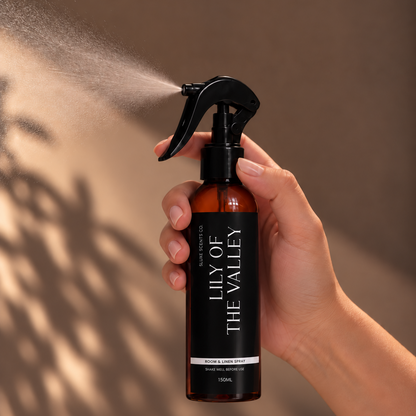 A hand holding a spray bottle of Lily of the Valley room spray, with the nozzle spraying a fine mist. The bottle is amber brown with a black spray head and a black label with white text.