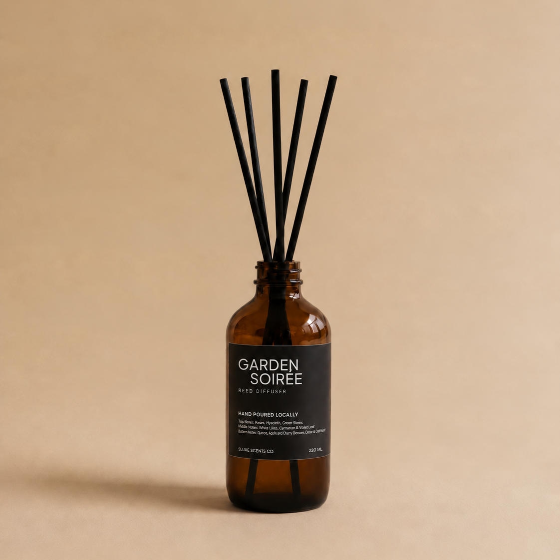 An amber glass reed diffuser bottle with a black label reading 'GARDEN SOIRÉE' and several black reed sticks, centered on a warm beige background.