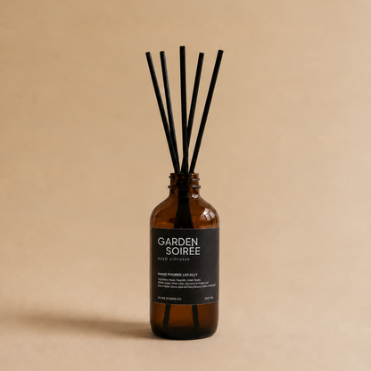 An amber glass reed diffuser bottle with a black label reading 'GARDEN SOIRÉE' and several black reed sticks, centered on a warm beige background.