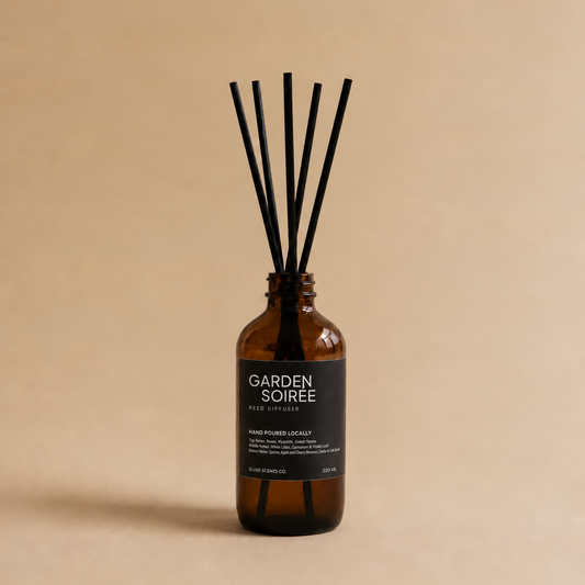 An amber glass reed diffuser bottle with a black label reading 'GARDEN SOIRÉE' and several black reed sticks, centered on a warm beige background.