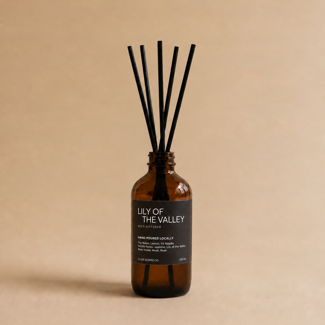 An amber glass reed diffuser bottle with a black label reading 'LILY OF THE VALLEY' and six black reeds, set against a solid beige background.