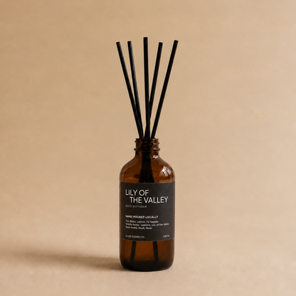 An amber glass reed diffuser bottle with a black label reading 'LILY OF THE VALLEY' and six black reeds, set against a solid beige background.