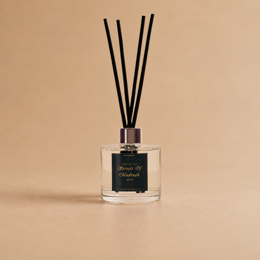 A reed diffuser bottle with a clear glass body, black reeds, and a black label with gold lettering, set against a solid beige background.