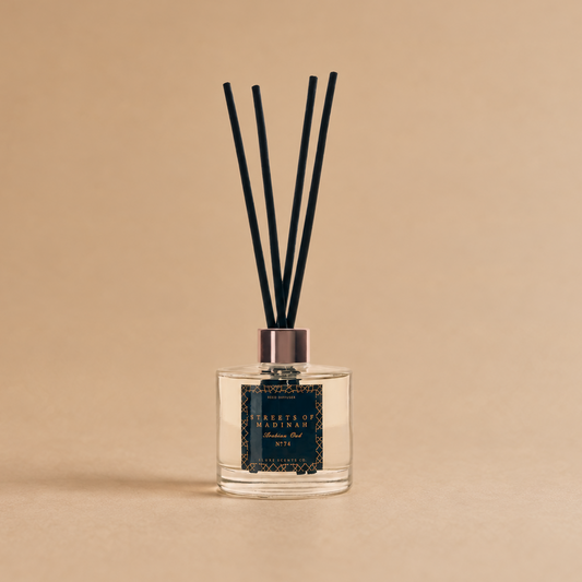 A reed diffuser bottle with a clear glass base, dark liquid, a patterned navy label, and four black reeds, centered on a warm beige background.